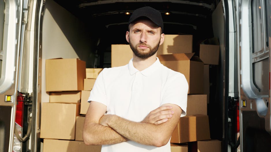 A young man with a beard and serious expression, wearing a black cap and a white polo shirt, stands inside the open rear doors of a moving van. Behind him, the van is filled with neatly stacked cardbo