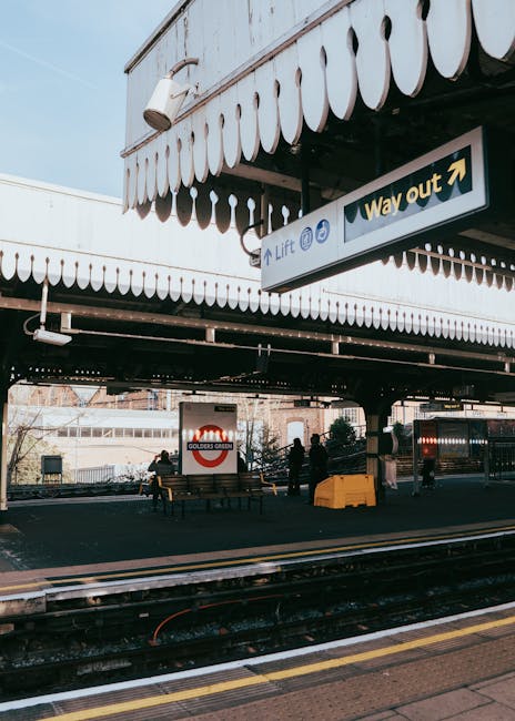 The image shows the exterior of Golders Green underground station with several passengers waiting on the platform, some sitting on wooden benches and others standing, beneath a large overhanging struc